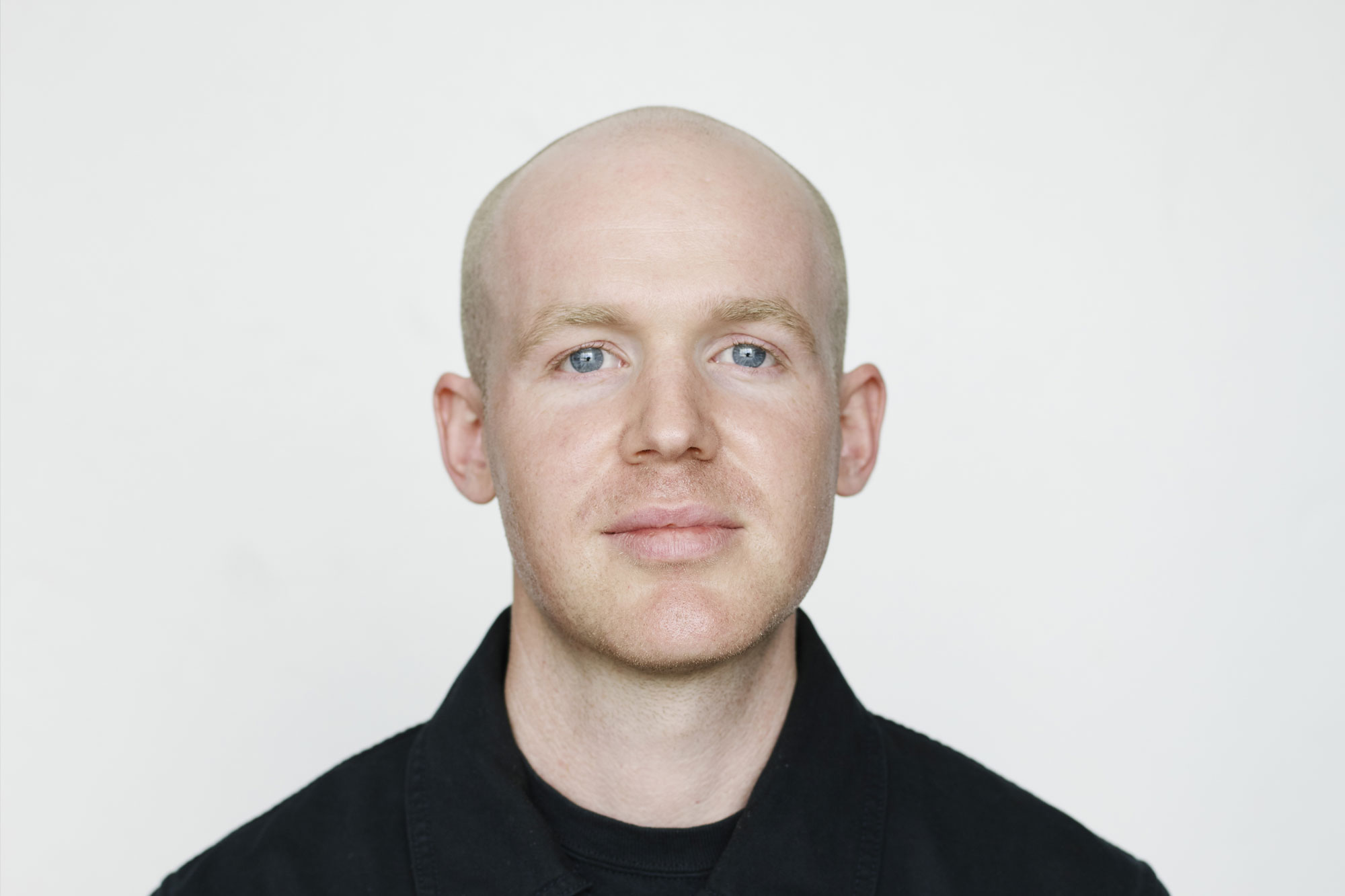 A person with closely-cropped fine hair on their crown and no hair on the top of their head, blue eyes, white skin. They are wearing a black button-up shirt and are sitting in front of a white wall.