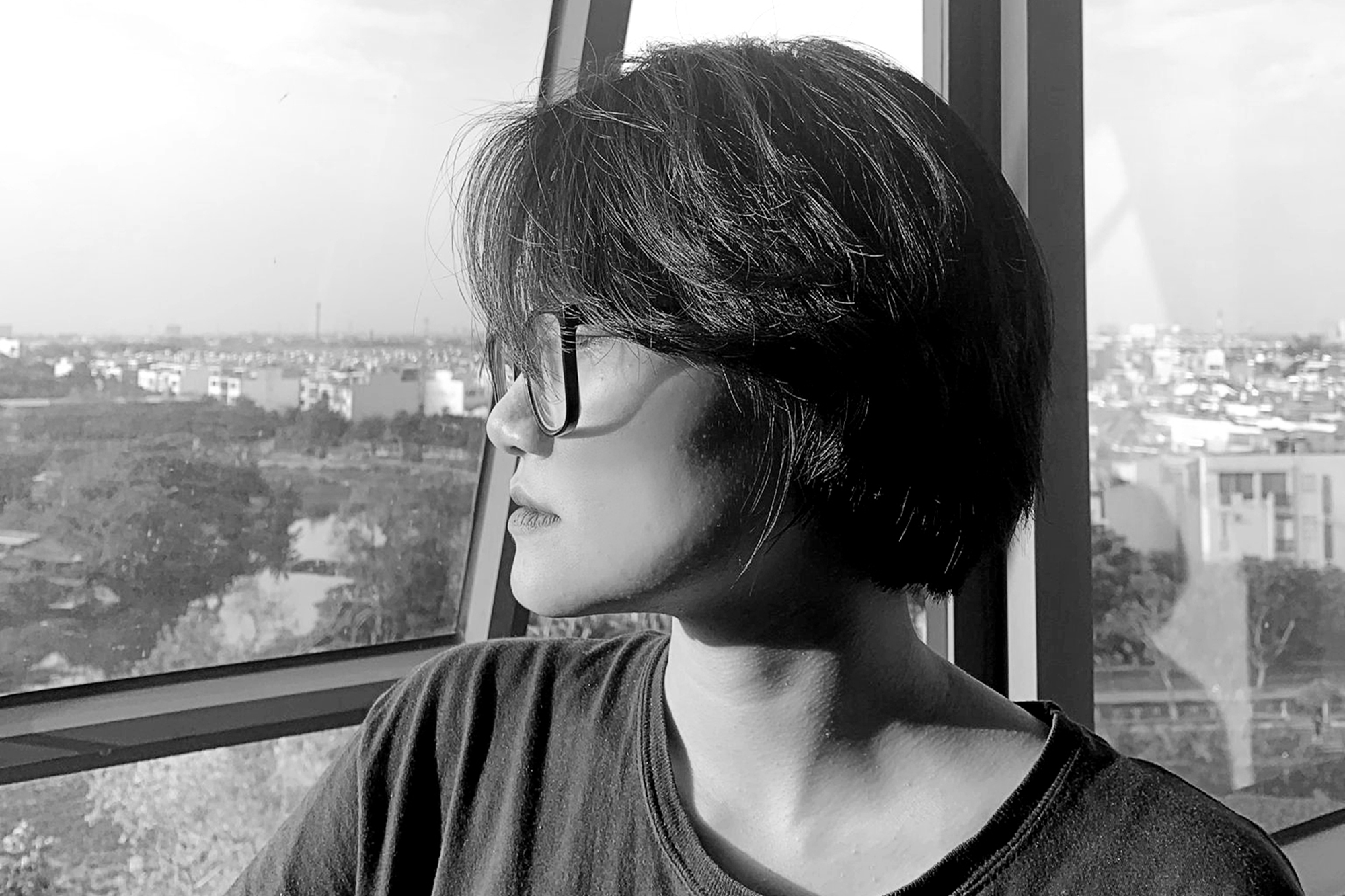 A person with medium-length hair cut in a layered bob with dark framed glasses and wearing a faded black t-shirt. They are looking out the window and in the distant background you can see trees and a city
