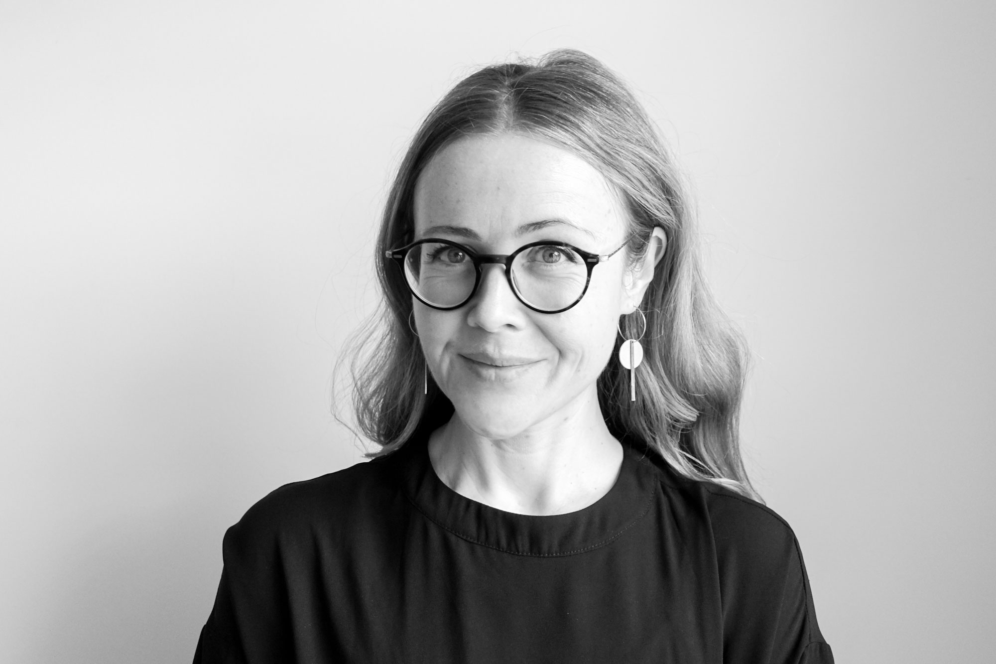 A black and white photo of a person with long light-coloured hair, light skin and eyes. They are wearing round glasses with a thin dark frame, dangling earrings in geometric shapes, and a dark-coloured shirt. They're smiling at the camera person and sitting in front of a light-coloured wall.