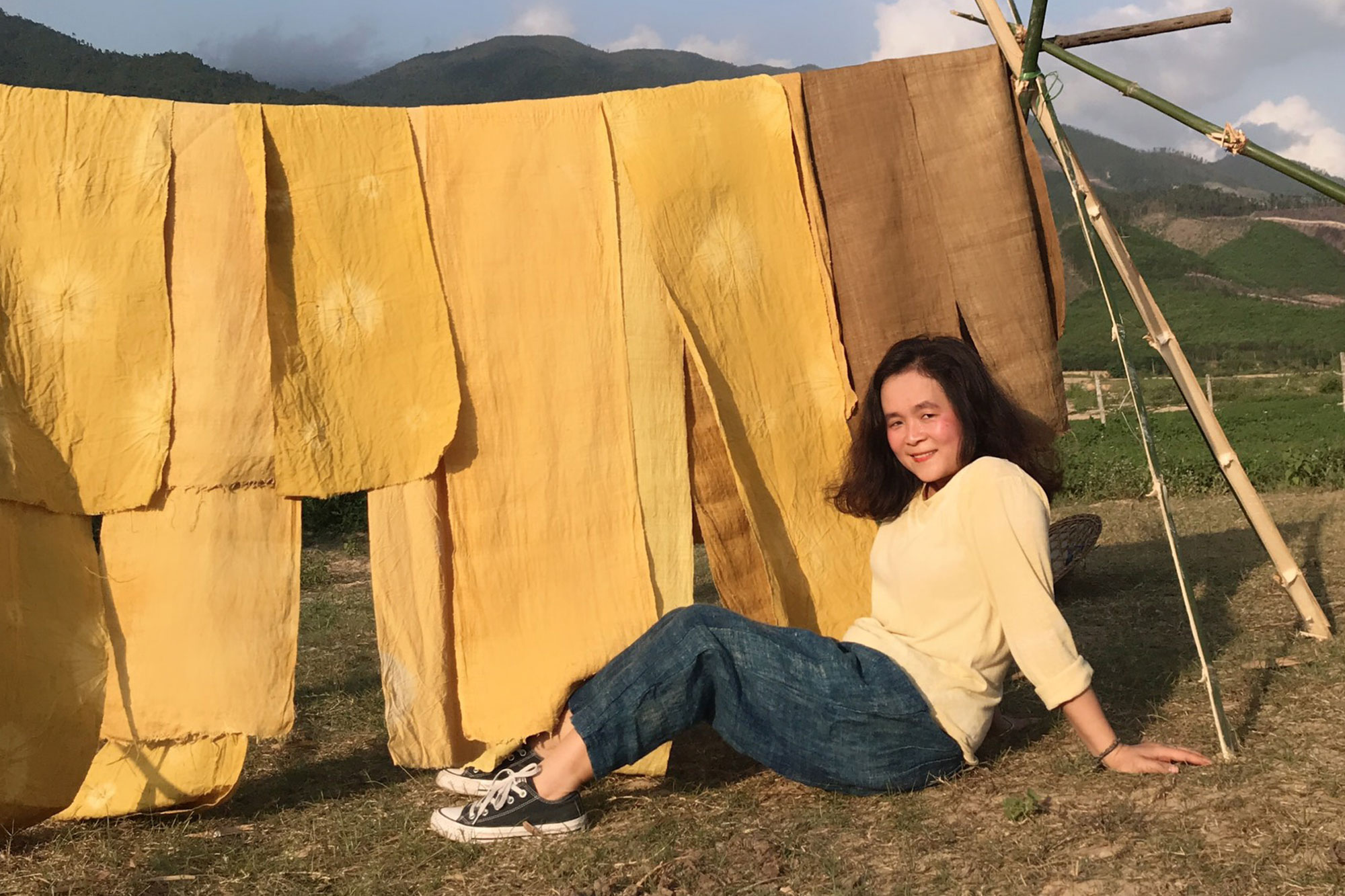A person sitting on grass with mountains and blue sky in the background. The're sitting in front of some yellow dyed fabric that is hanging to dry. The person is smiling, has long, dark, currly hair and is wearing a long sleeved light yellow top, jeans and some black and white sneakers.