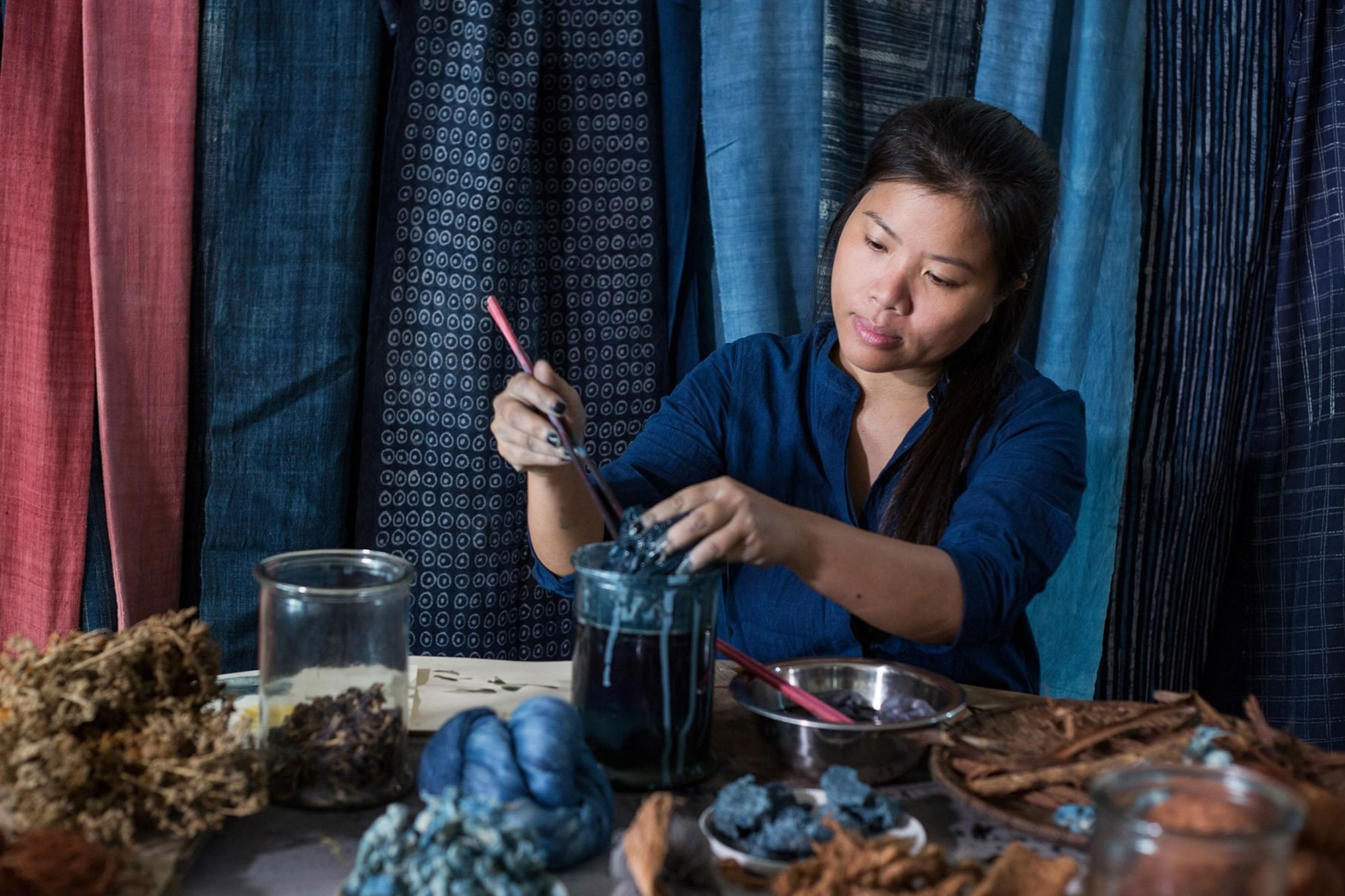 A person with brown skin, dark eyes and long dark hair sitting in front of a blue and red curtain and using a paintbrush to dip into a cylindrical beaker filled with blue liquid. There is another  beaker ion the table and some natural objects. Ther person is wearing a blue shirt.